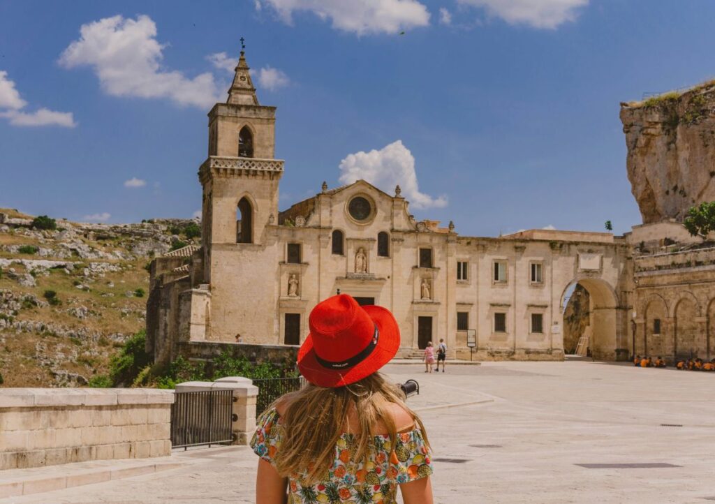 A blonde girl wearing a red a hat and flowery dress walking towards an ancient church in Italy